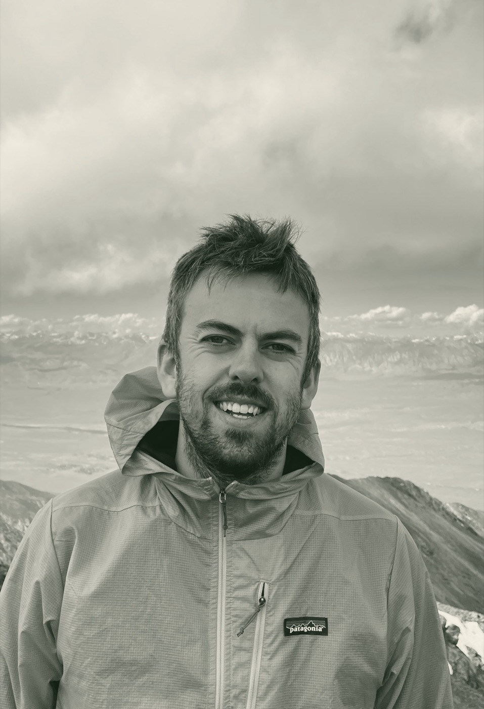 Rob Price, smiling, on a mountain summit under a cloudy sky